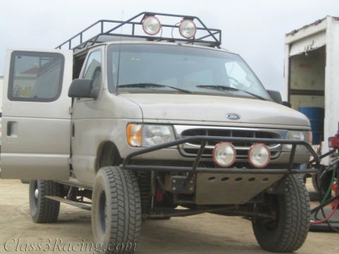 1990 Ford Bronco - Front view of a dusty silver Ford van lifted on off-road tires with a tube bumper, auxiliary lights and roof rack on a dirt lot.
