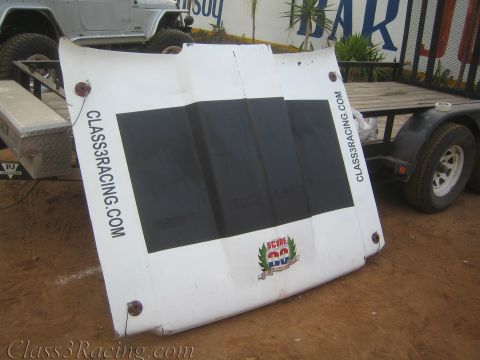 1990 Ford Bronco - white fiberglass race-truck hood marked CLASS3RACING.COM with a black center panel and SCORE logo leans against a utility trailer on dirt.