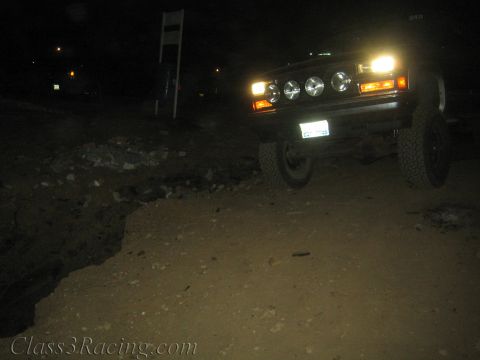 1990 Ford Bronco - lifted boxy SUV with roof-mounted lights illuminated crawls along a rocky, washed-out dirt road at night.
