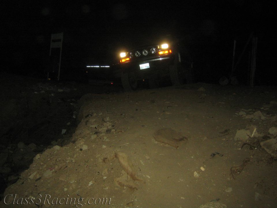 Lifted vehicle with headlights and amber markers on in darkness behind a dirt berm
