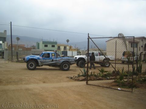 1990 Ford Bronco - blue tube-chassis race truck sits beside white chase trucks behind an open chain-link gate in a dusty Baja-style lot.