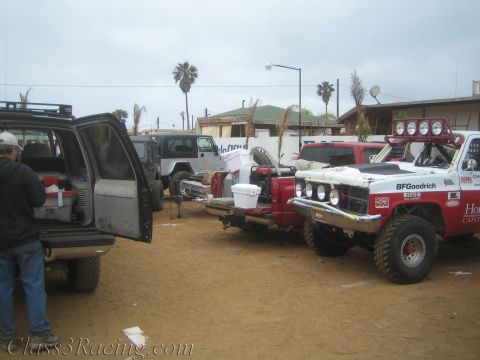 1990 Ford Bronco - Race and chase vehicles crowd a dusty pit area, including a red-and-white off-road truck with BFGoodrich livery and a Jeep.