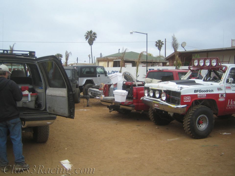 Off-road vehicles parked in a dusty pit area, including a red-and-white race truck with BFGoodrich livery