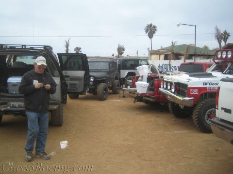1990 Ford Bronco - man in a cap stands by an open SUV door in a dusty lot lined with off-road race and chase trucks under overcast sky.