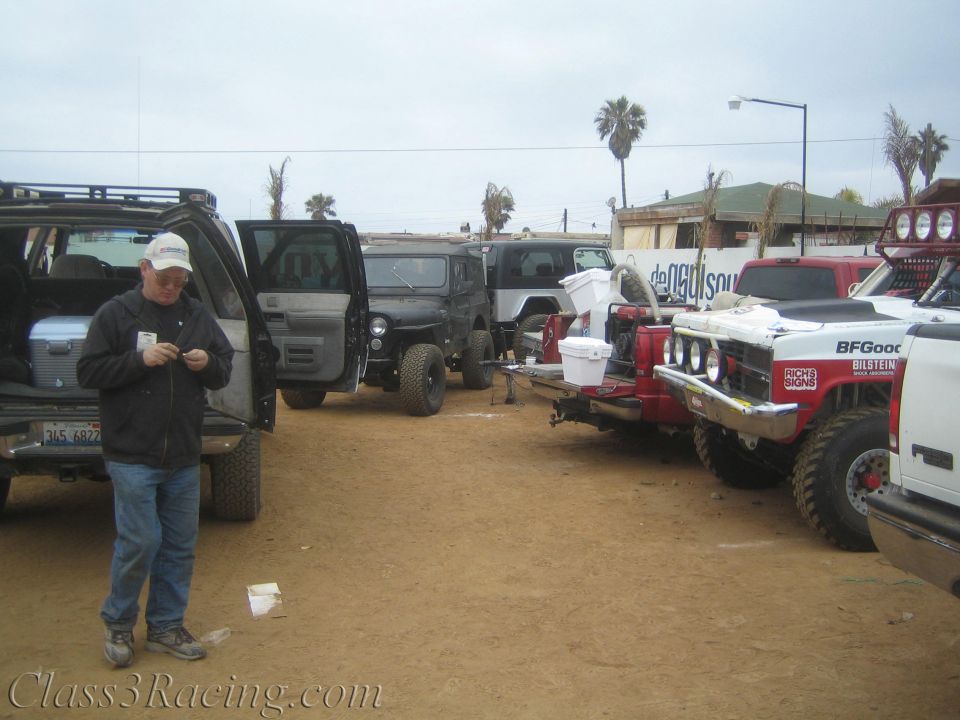 Man in a cap standing beside open SUV doors in a dusty staging lot full of off-road trucks