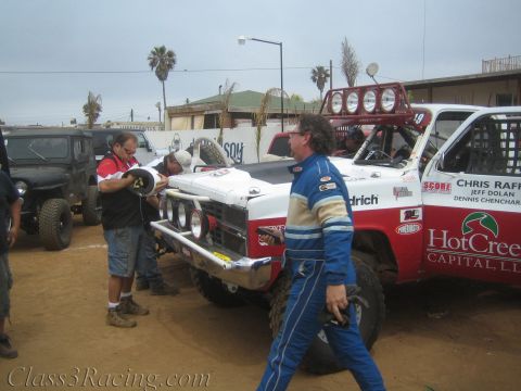 1990 Ford Bronco - Crew members service a red-and-white SCORE off-road race truck while a driver in a blue racing suit walks past in a dusty lot.