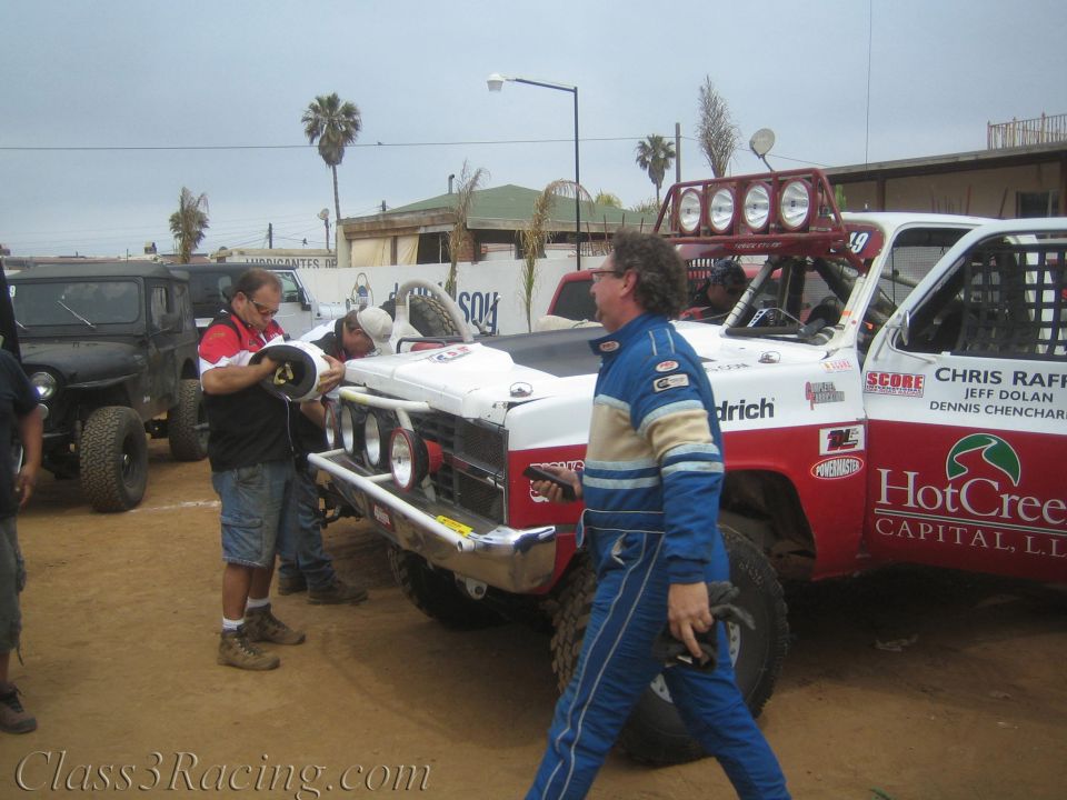 Crew working on a red-and-white off-road race truck as a driver in blue suit walks past