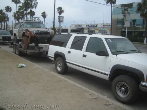 1990 Ford Bronco - white Chevrolet Suburban tows a flatbed trailer loaded with a white lifted Ford Bronco past a tire shop on a city street.