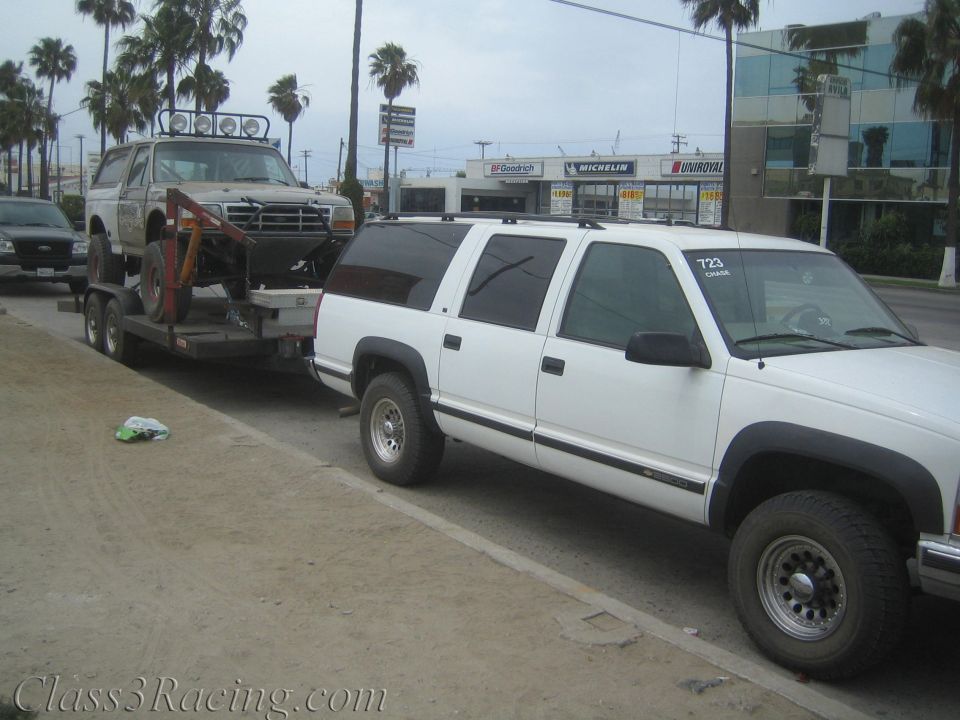 White Chevrolet Suburban towing a trailer carrying a white lifted Ford Bronco on a street