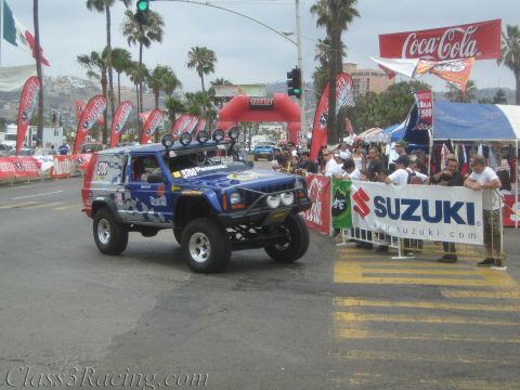 1990 Ford Bronco - blue lifted Jeep Cherokee race vehicle rolls past a crowd at a street start line flanked by Coca-Cola and Suzuki banners.