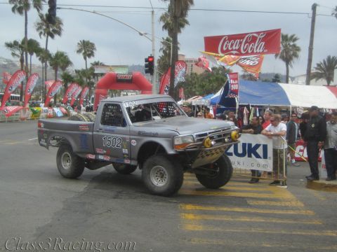 1990 Ford Bronco - Silver Ford prerunner-style race truck No. 1502 stages at a palm-lined street start line beneath a Coca-Cola banner.