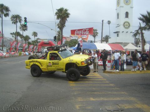 1990 Ford Bronco - Yellow lifted mini race truck No. 1549 sits at a palm-lined street intersection with a white clock tower in the background.