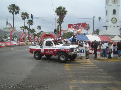 1990 Ford Bronco - White Chevy off-road race truck No. 349 with HotCreek Capital livery rolls past spectators at a palm-lined street start line.