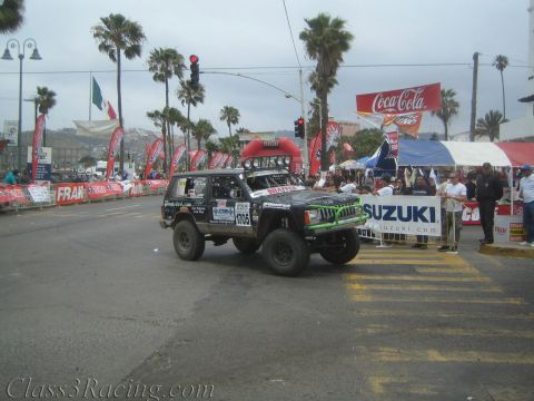 1990 Ford Bronco - black-and-green lifted Jeep Cherokee race vehicle No. 1705 crosses a street start line beside a Suzuki banner.