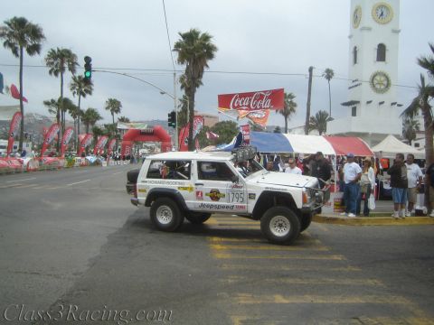 1990 Ford Bronco - White lifted Jeep Cherokee No. 1795 with Jeepspeed livery rolls past spectators at a street start line by a clock tower.