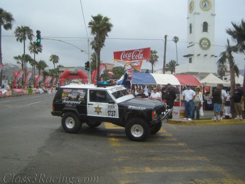 1990 Ford Bronco - black-and-white Jeep Cherokee race vehicle No. 1737 with sheriff-style graphics stages at a palm-lined street start line.