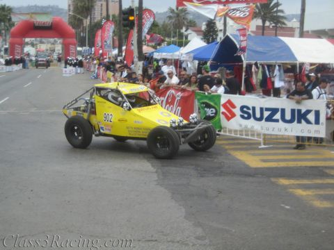 1990 Ford Bronco - yellow single-seat off-road race buggy No. 902 rolls past spectators at a street start line beside a Suzuki banner.