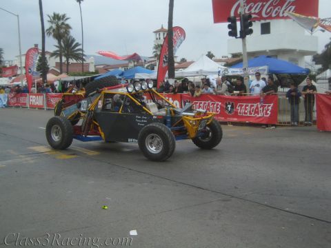 1990 Ford Bronco - yellow tube-frame off-road buggy carrying a spare tire drives down a street past Tecate and Coca-Cola banners.