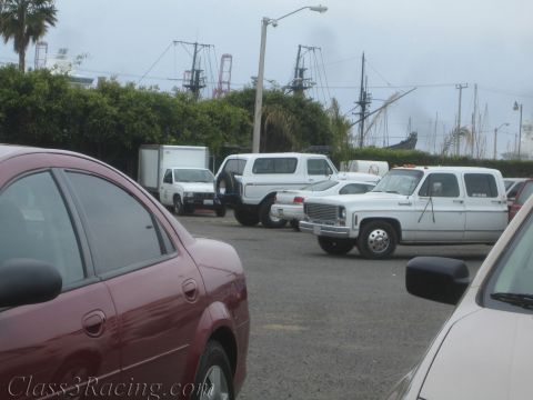 1990 Ford Bronco - Cars and white work trucks fill a waterfront parking lot, with tall sailing-ship masts and a large gray ship beyond the hedges.