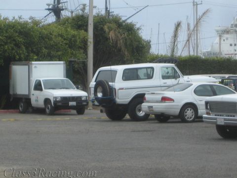 1990 Ford Bronco - white Ford Bronco with rear spare tire sits among a box truck, cars, and a white pickup in a harborside lot with ship masts behind.
