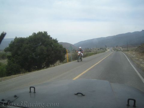 1990 Ford Bronco - View over a vehicle hood of a helmeted rider on a dirt bike riding toward the camera on a rural two-lane mountain road.