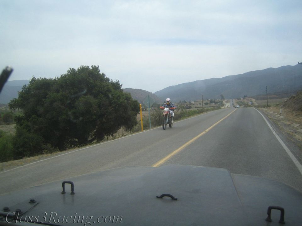 Helmeted rider on a dirt bike approaching on a rural two-lane road seen from a vehicle hood