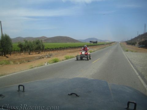 1990 Ford Bronco - rider on a red ATV quad travels down a two-lane desert highway past vineyard rows and distant hills.
