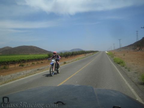 1990 Ford Bronco - helmeted rider on a dirt motorcycle heads down an empty two-lane road bordered by green vineyards and dry hills.