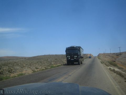 1990 Ford Bronco - dark off-road truck travels along a remote desert highway flanked by sparse scrub and low hills.