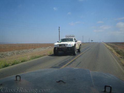 1990 Ford Bronco - white off-road truck fitted with a front roof light bar sits along a two-lane desert highway under clear skies.