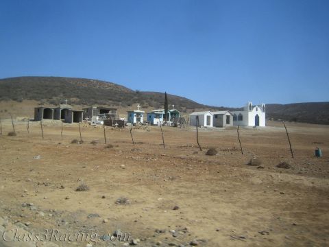 1990 Ford Bronco - small settlement of houses and a white peaked chapel stands on dry desert ground below brown hills.