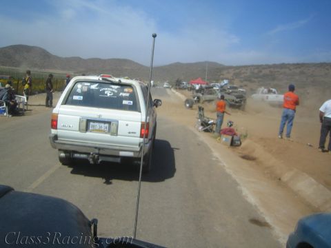 1990 Ford Bronco - white SUV with a tall whip antenna sits roadside amid dusty off-road race pits, spectators and vehicles.