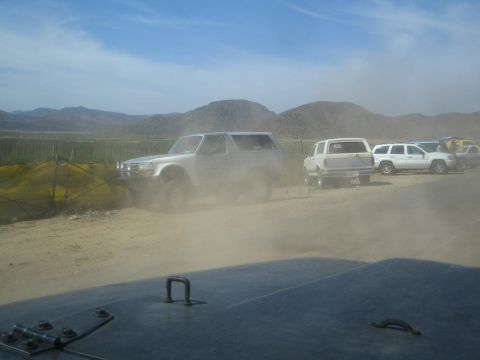 1990 Ford Bronco - silver lifted Bronco-style SUV and other trucks sit in a dusty desert staging area near a fence.