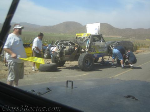 1990 Ford Bronco - pit crew works on a yellow-and-silver number 102 off-road race buggy stopped along a desert roadside.