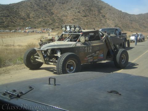 1990 Ford Bronco - tan number 112 tube-frame off-road race buggy with roof light pods rolls along a desert road past spectators.