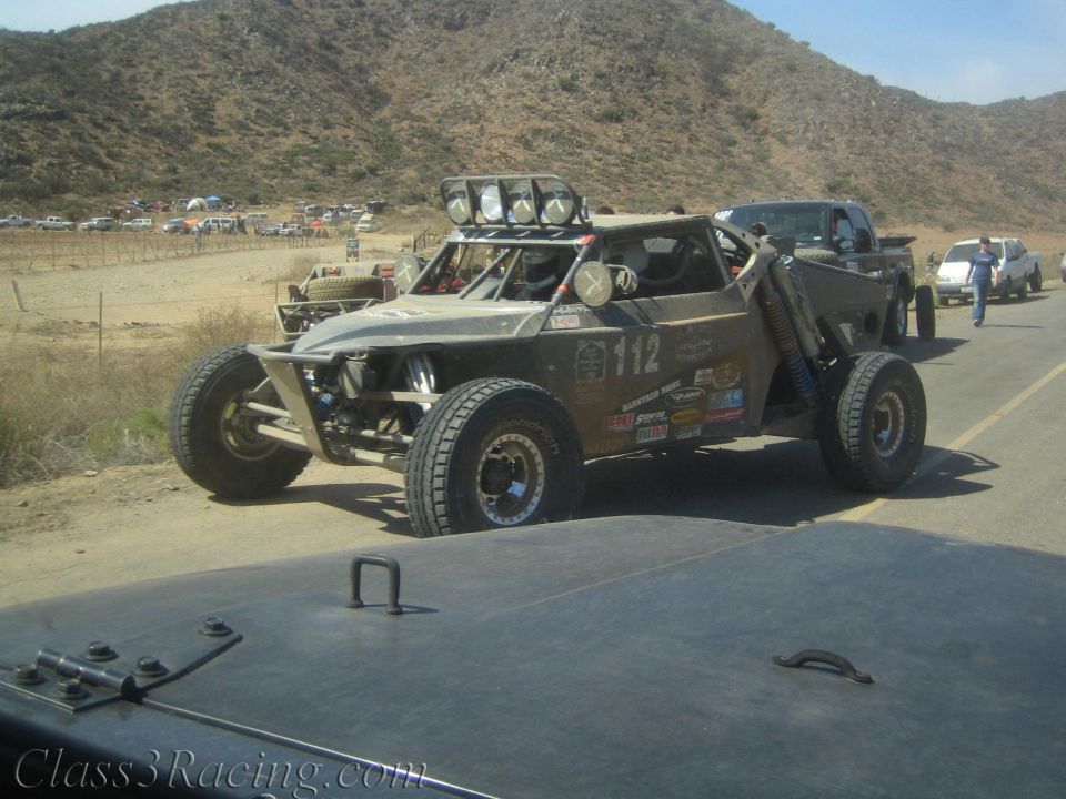 Number 112 tan off-road race buggy with roof light pods on a desert road