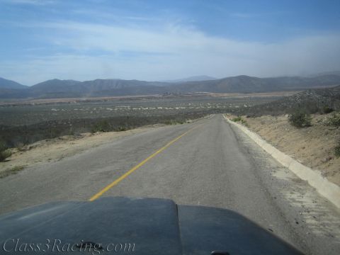 1990 Ford Bronco - Seen over a dark vehicle hood, an empty two-lane highway curves down into a broad arid desert valley.