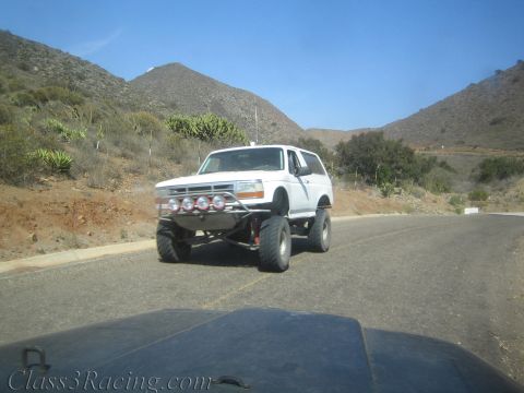 1990 Ford Bronco - white lifted Bronco-style SUV with a front bumper light bar drives a winding road through dry cactus-dotted hills.