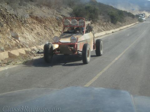 1990 Ford Bronco - weathered open-frame off-road buggy with roof lights drives a paved road cut into a rocky hillside.