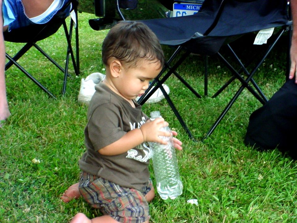 Toddler kneeling on grass, holding a plastic water bottle. A Michigan license plate is visible in the background