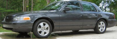 1985 Ford LTD Crown Victoria - A dark gray sedan is parked outdoors on a gravel surface, with trees visible behind it.
