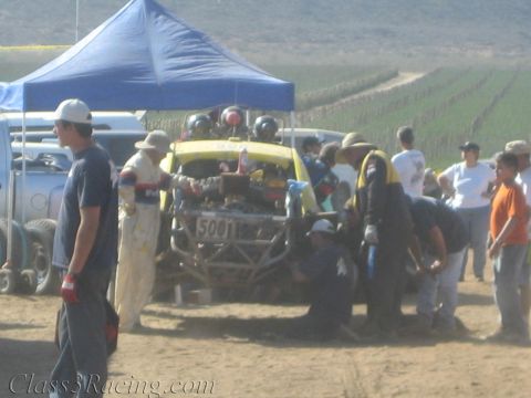 1990 Ford Bronco - crew tends a yellow tube-frame off-road buggy in a dusty desert pit area beside canopies and field rows.