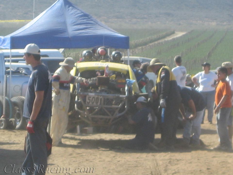 Crew works on a yellow off-road buggy at a dusty desert pit under canopies