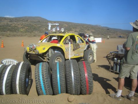 1990 Ford Bronco - yellow off-road race buggy sits at a desert pit beside rows of colorfully striped spare tires and crew.