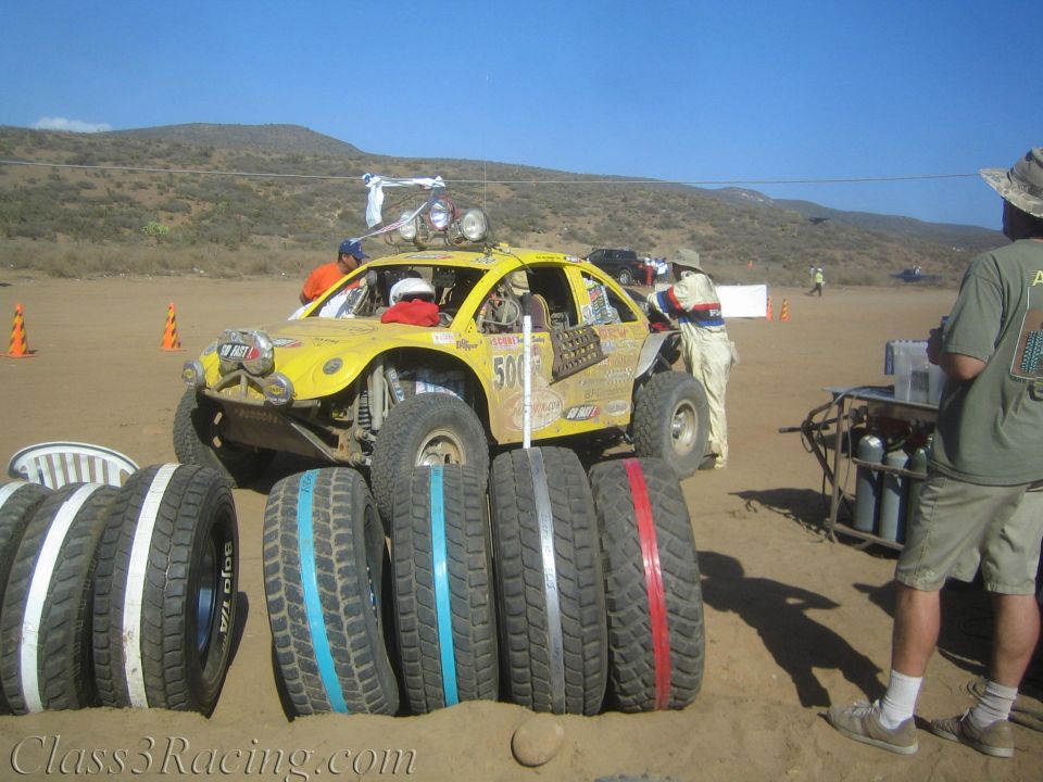 Yellow off-road buggy and rows of spare tires at a dusty desert pit