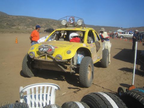 1990 Ford Bronco - yellow tube-frame off-road race buggy with roof light pods faces forward in a dusty desert pit area.