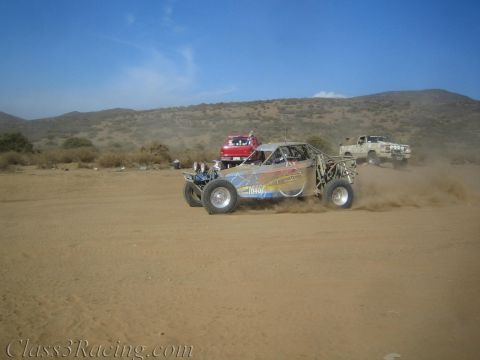 1990 Ford Bronco - off-road race buggy throws up dust crossing a desert flat as a red and white chase truck follow behind.