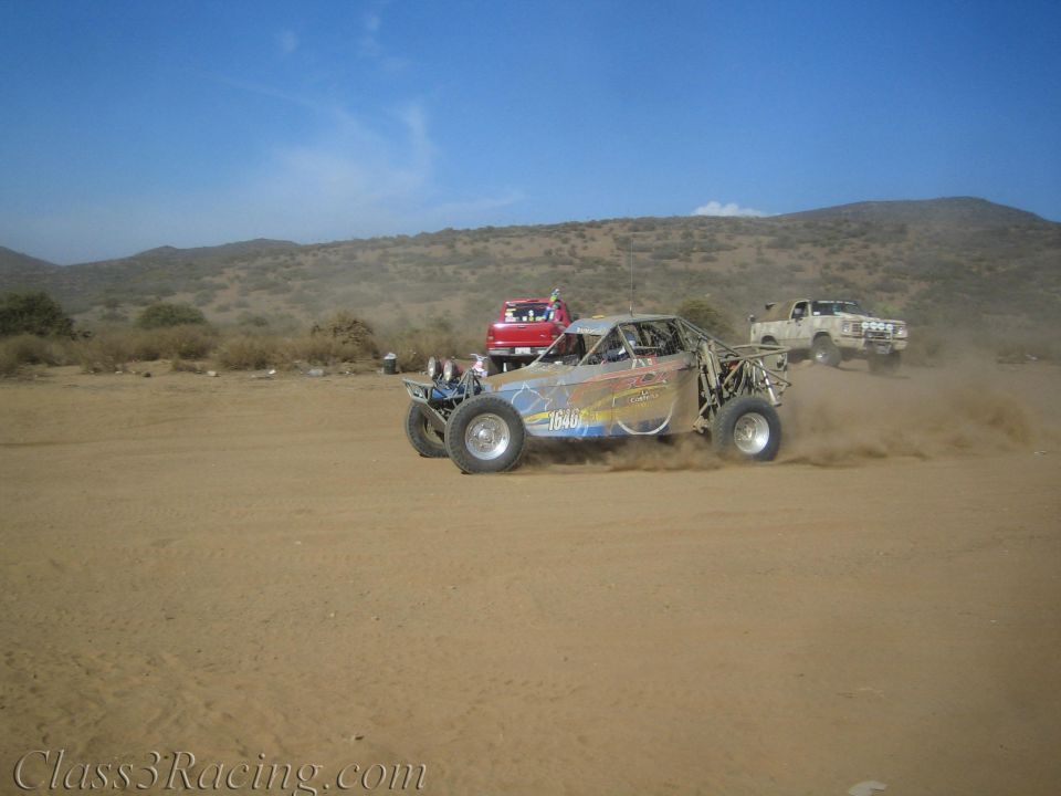 Off-road buggy kicking up dust on a desert flat with chase trucks behind