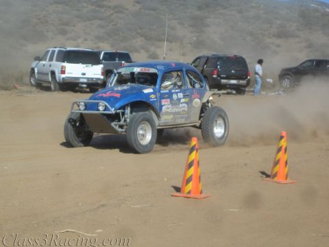 1990 Ford Bronco - blue off-road race buggy navigates between orange-striped cones on a dusty desert flat near parked trucks.