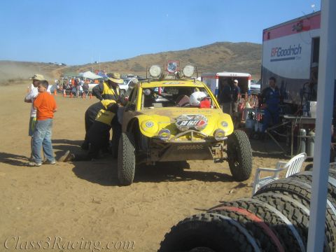 1990 Ford Bronco - Crew members work on a yellow off-road buggy in a dusty desert pit beside a BFGoodrich trailer and spare tires.